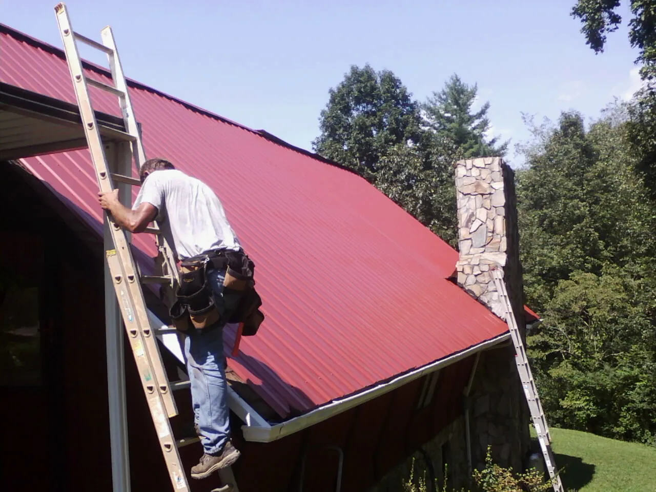 Roofer on a ladder beside a red metal roof and stone chimney — Soffit & Fascia Repair by Roofing Brothers in Wrightsville